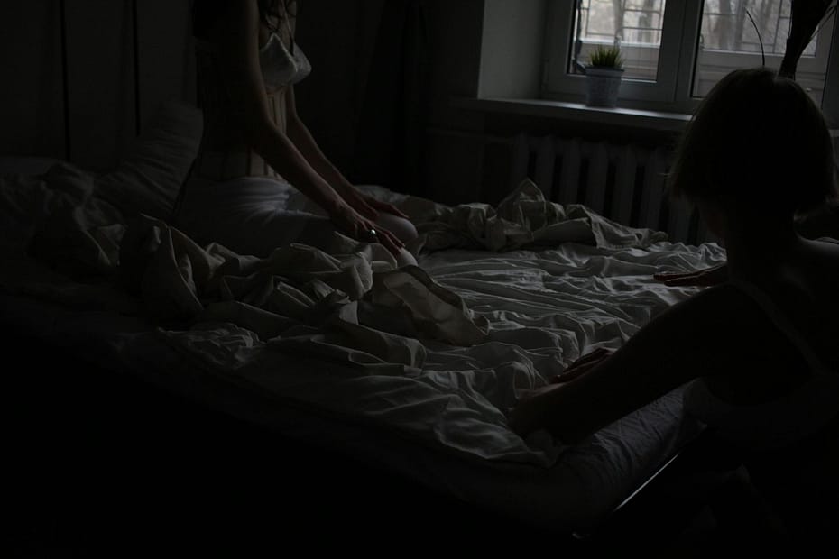 A dark room with two women standing on the corners of a messy bed trying to manifest while you sleep.